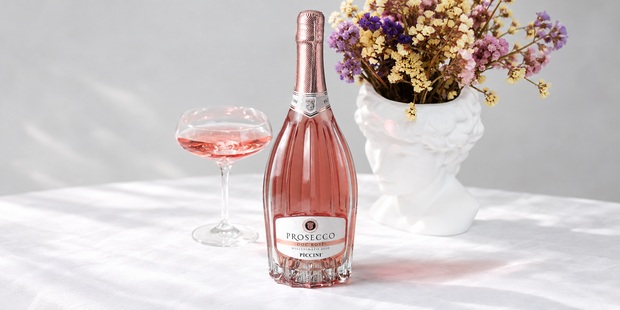 Lahev Prosecca Rosé Piccini se sklenkou a vázou s květinami.